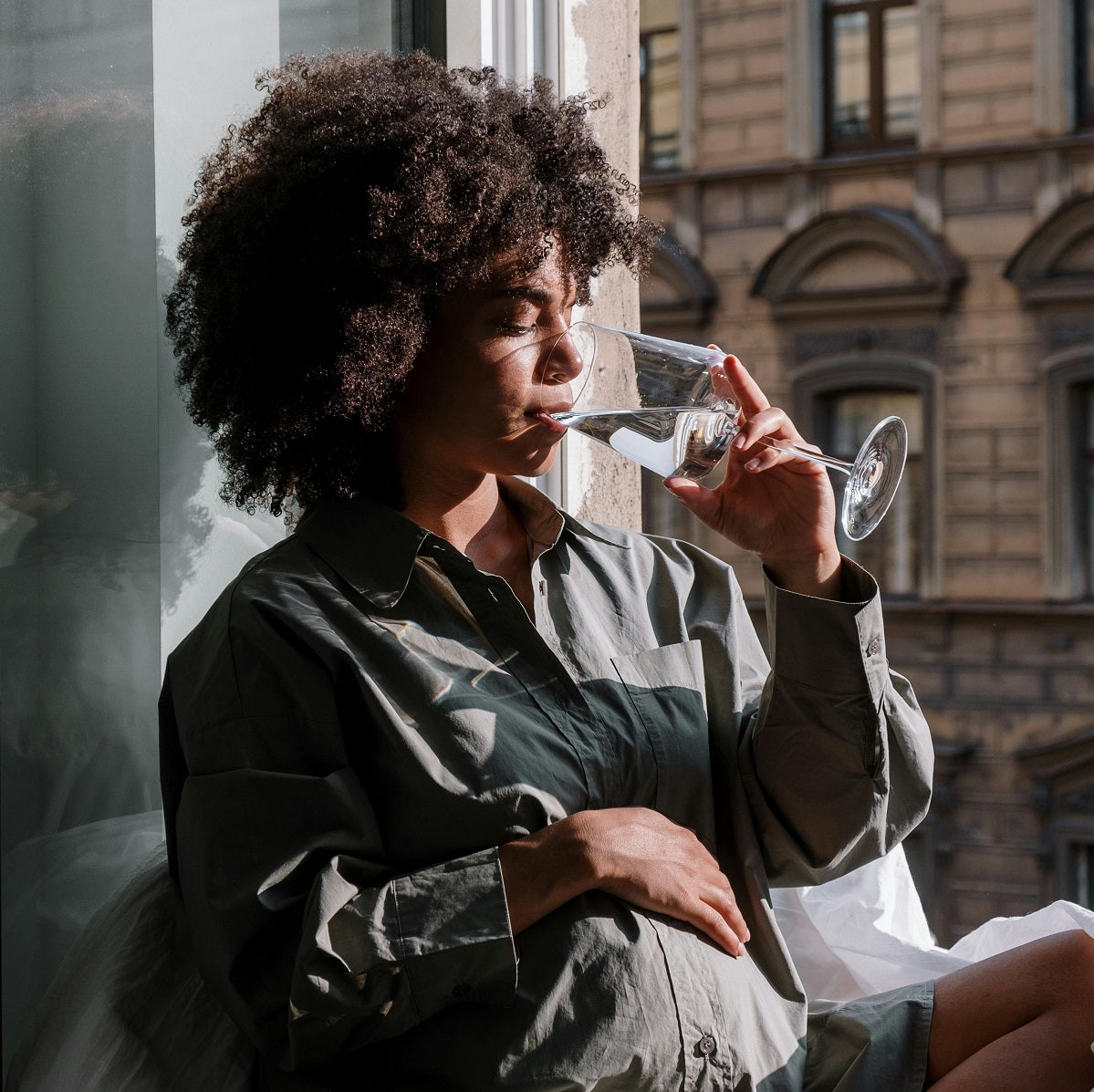 A woman sits by a sunlit window drinking water from a glass while resting a hand on her pregnant belly.